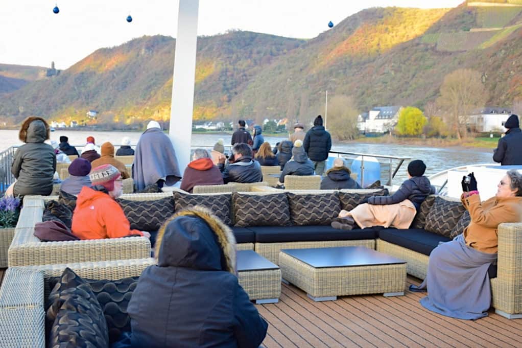 Passengers on deck wearing coats and hats along the Rhine river.